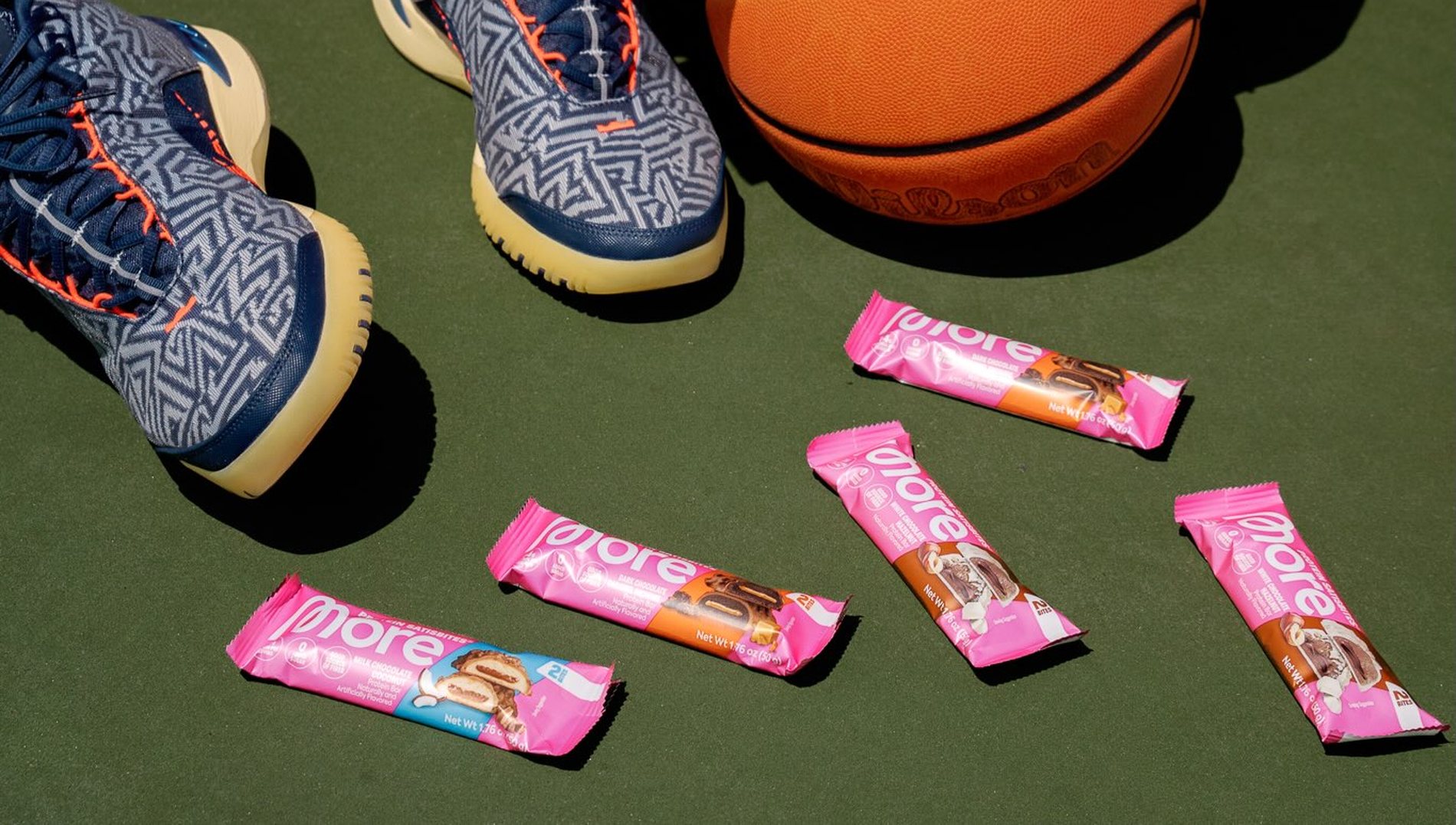 Five More Protein Satisbites packs laid out on a green outdoor court beside a basketball.